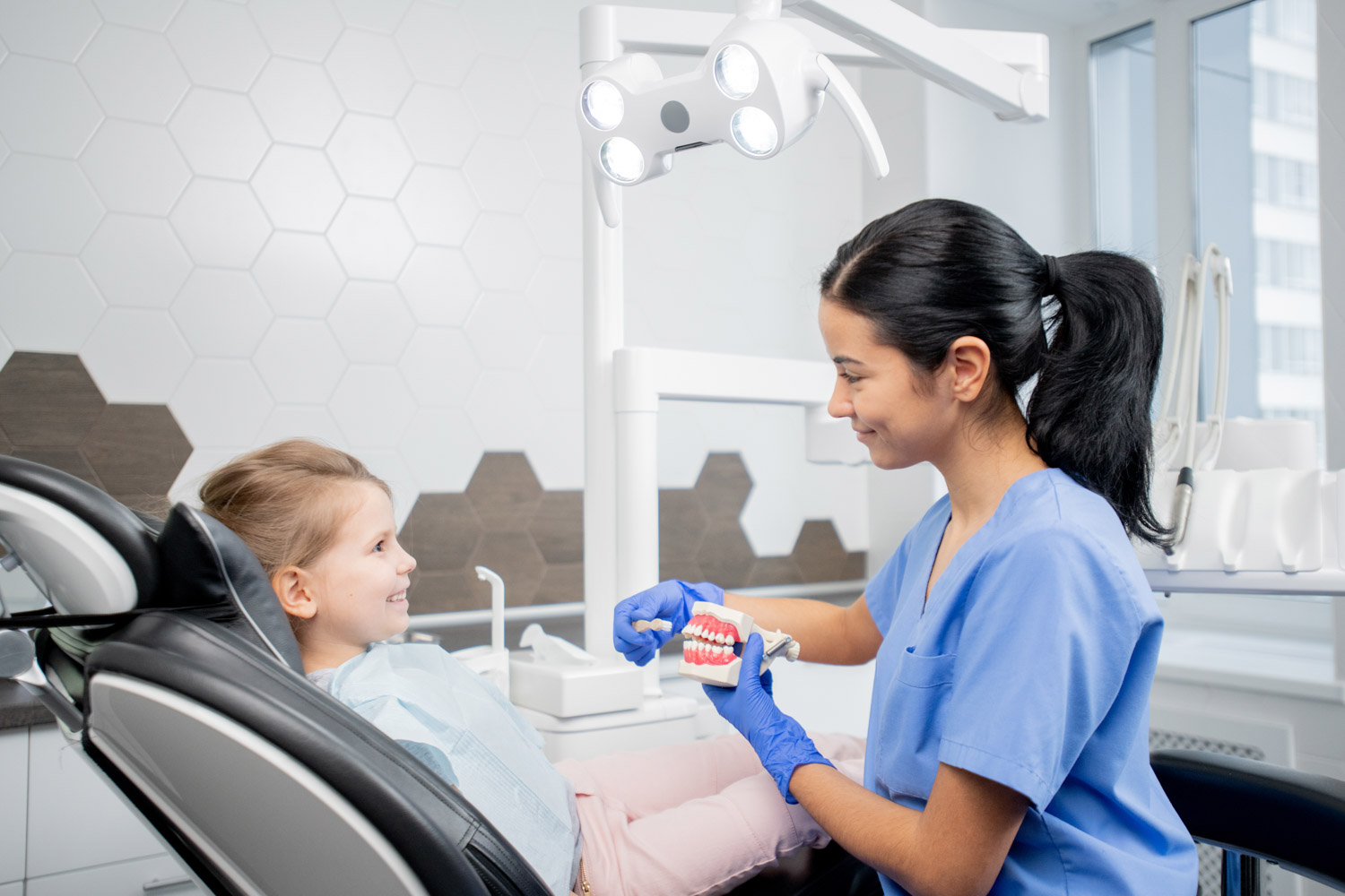Pretty young dentist in blue uniform showing little patient how to brush teeth